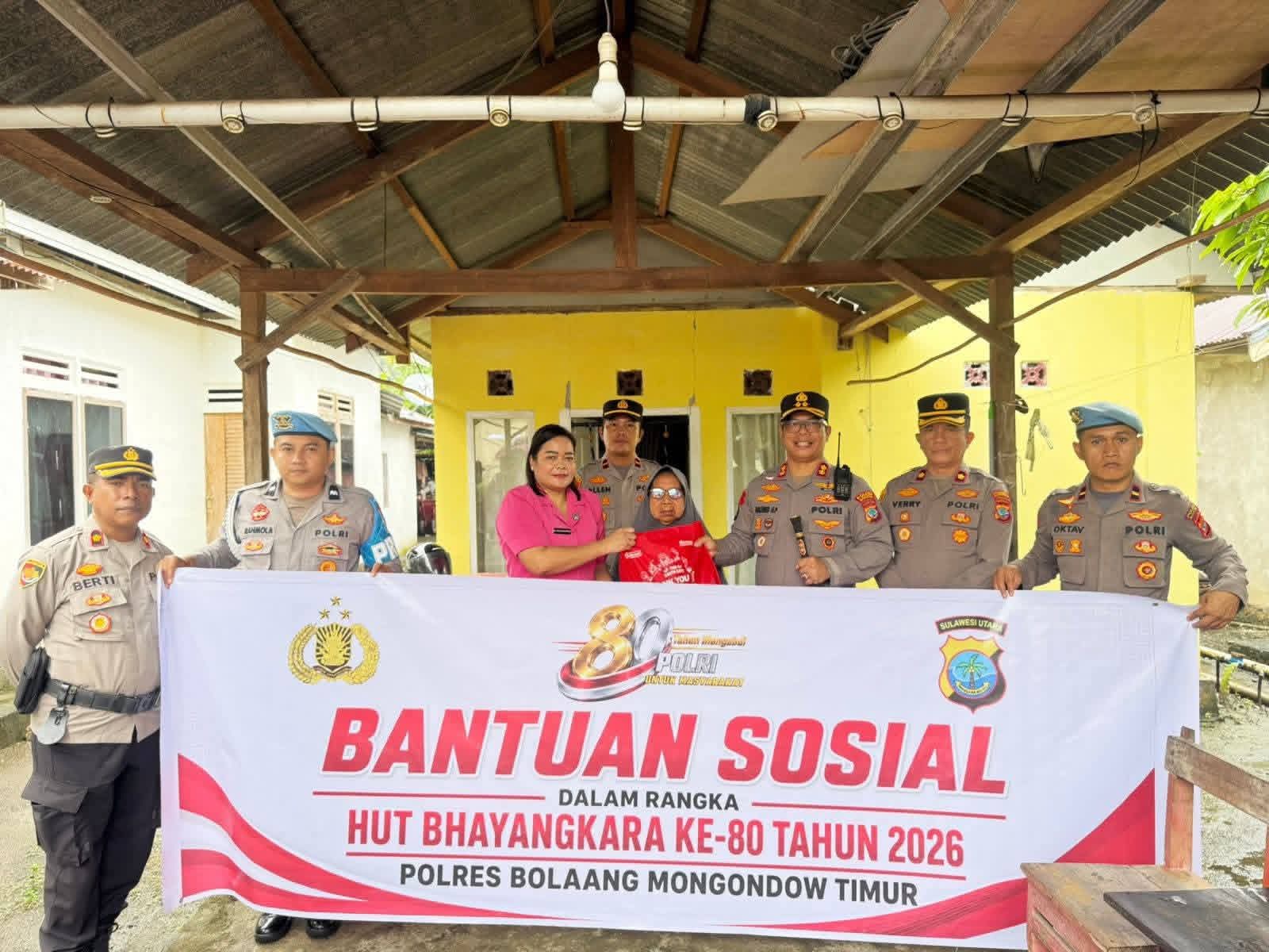 Sambut Hari Bhayangkara ke-80, Kapolres Boltim AKBP Golfried Hasiholan Pakpahan, S.H., M.Si. (ketiga dari kanan) bersama Ketua Bhayangkari Cabang Boltim Ny. Joice Golfried (keempat dari kanan) menyerahkan bantuan sosial berupa kemeja kepada seorang warga lansia (penerima manfaat) di Kecamatan Tutuyan. Penyerahan simbolis ini dilakukan di depan spanduk acara saat aparat kepolisian menyambangi warga secara door-to-door sebagai bentuk nyata kepedulian di tengah guyuran hujan.