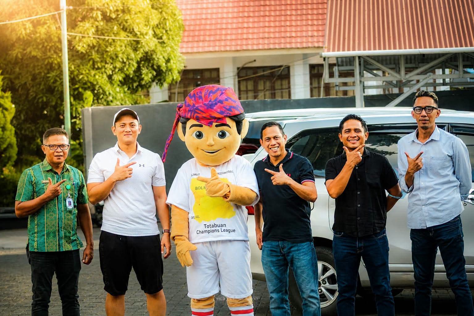 Ketua Askot PSSI Kotamobagu dengan kaos putih berdiri di samping maskot resmi Totabuan Champions League (TCL) 2026. Di sisi kanan, Wali Kota Kotamobagu dr. Weny Gaib mengenakan kaos hitam, bersama-sama menunjukkan dukungan atas peluncuran maskot “Bojo” yang akan memeriahkan turnamen di Stadion Nunuk Matali.